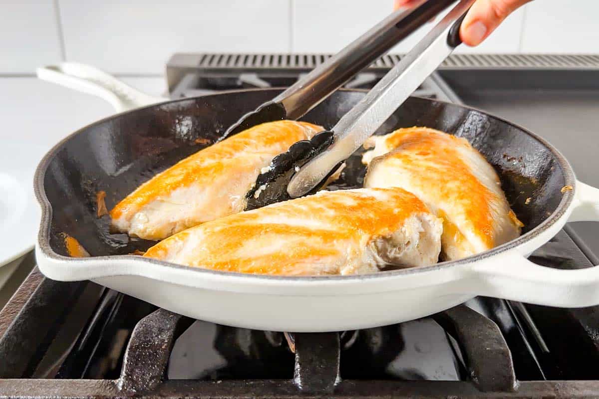 Searing the chicken breasts in a large white cast iron skillet turning a chicken breasts with a pair of tongs.