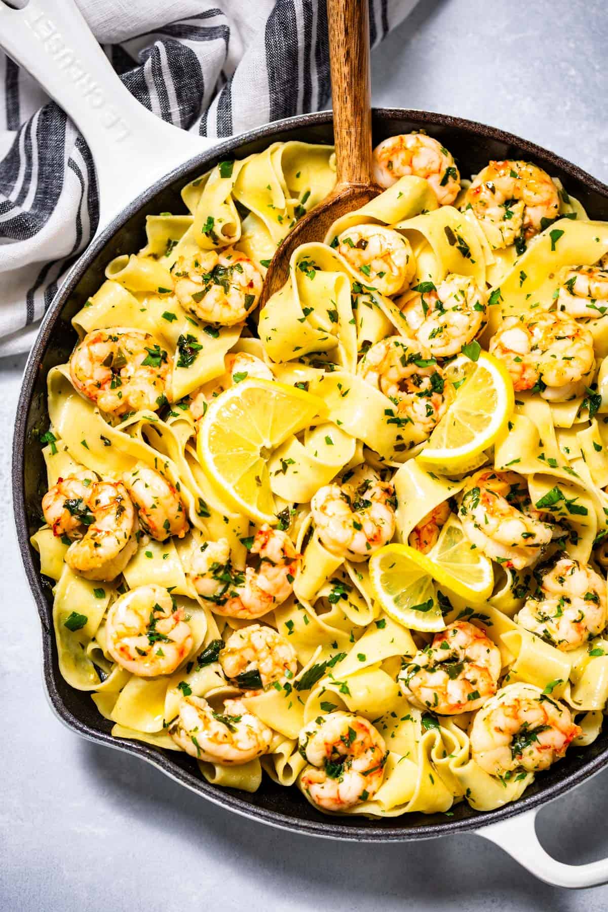 Garlic butter shrimp pasta in a large with skillet topped with lemon slices and chopped parsley and a wood spoon on the side.