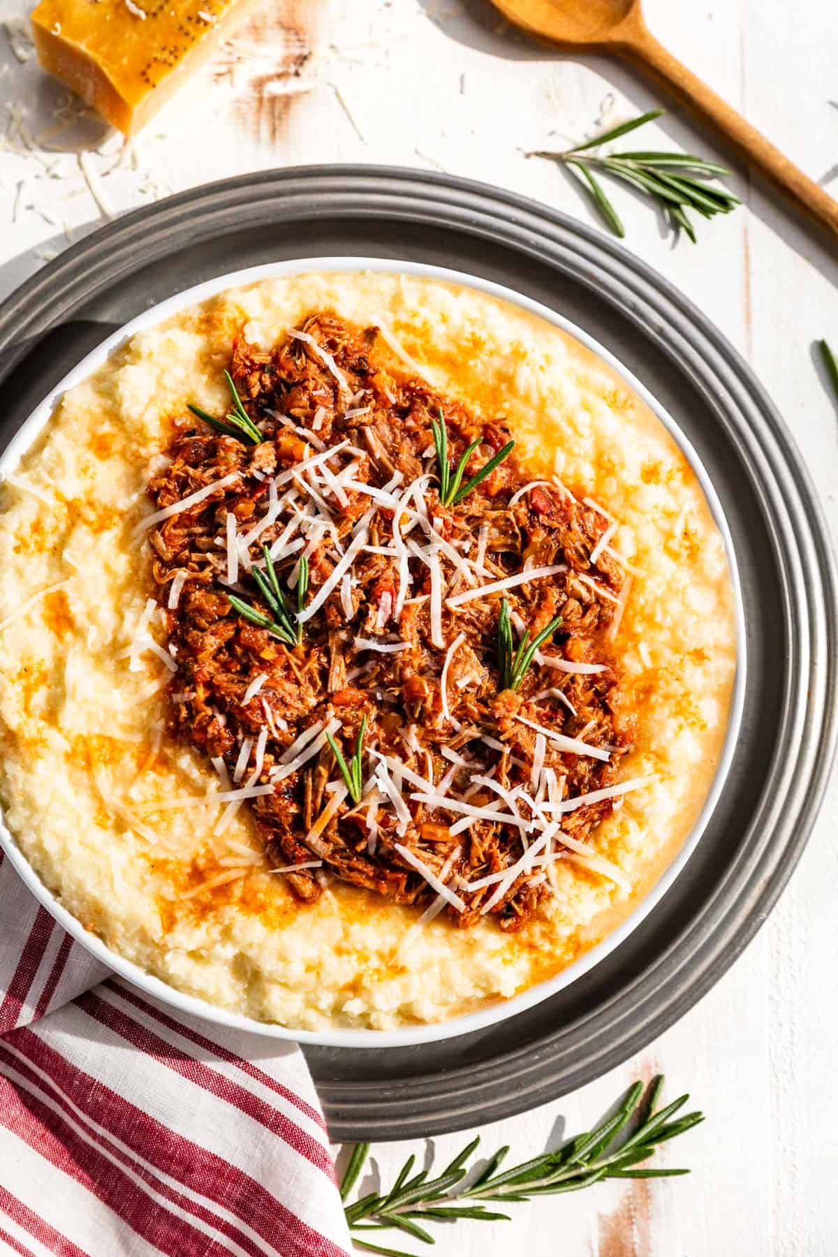 Pork ragu spooned over cauliflower polenta in a large white serving bowl topped with grated parmesan and garnished with rosemary sprigs.