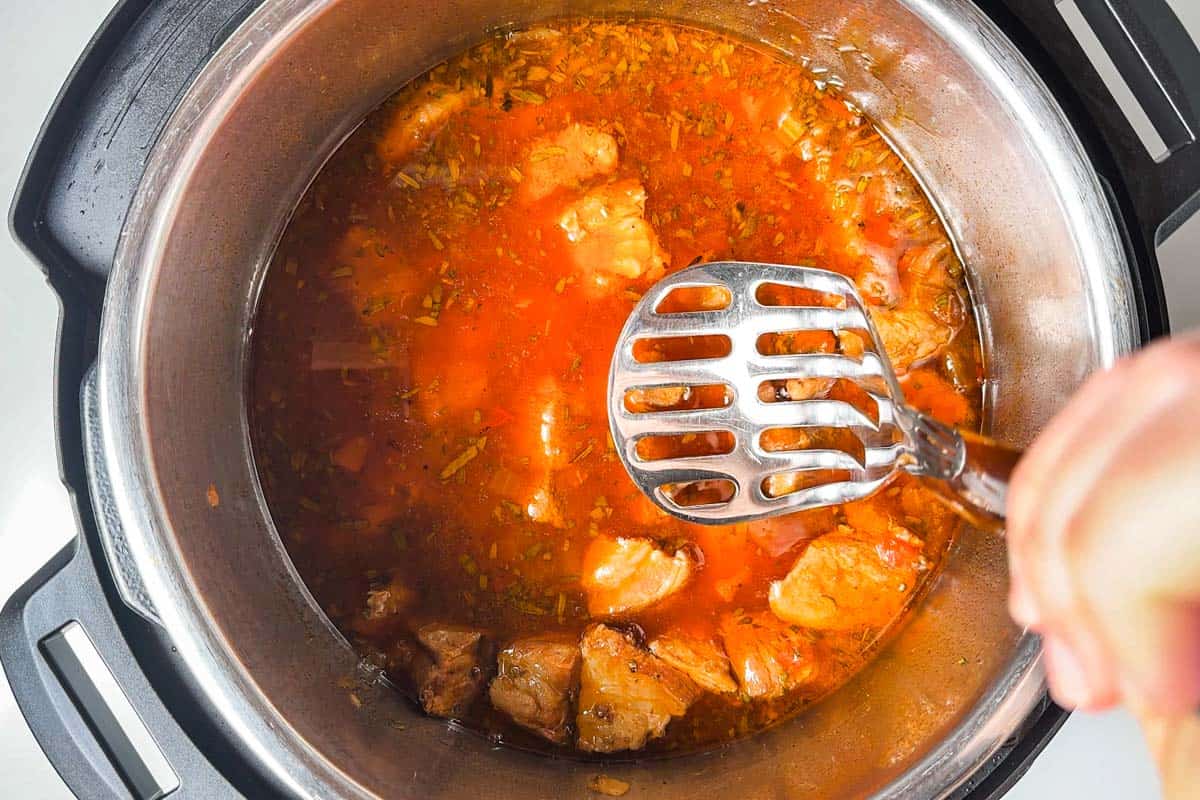Using a potato masher to break up the cooked pork shoulder cubes into shreds in the tomato broth in the metal bowl of the Instant Pot.