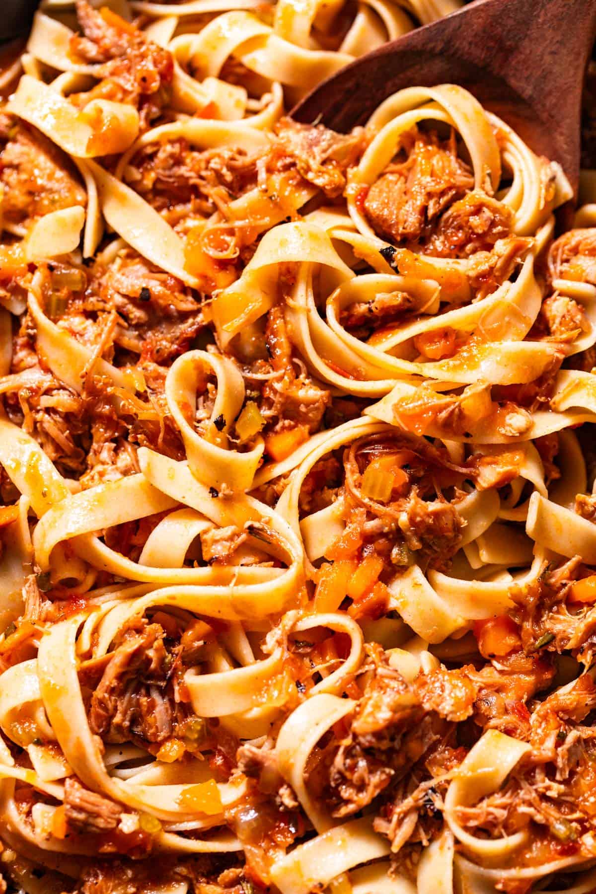 Close up shot of swirls of pasta tossed with pork ragu with a wood spoon in the noodles on the side of the bowl.
