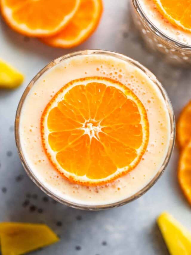 Two glasses of pineapple orange banana smoothie topped with thinly sliced oranges and pineapple tidbits around the glasses.