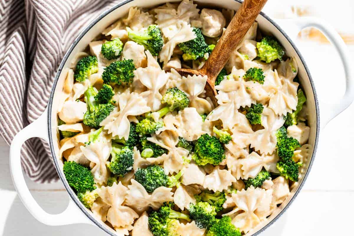 Chicken Broccoli Alfredo in a large white pot with a wood spoon in the pasta and a brown linen on the side.