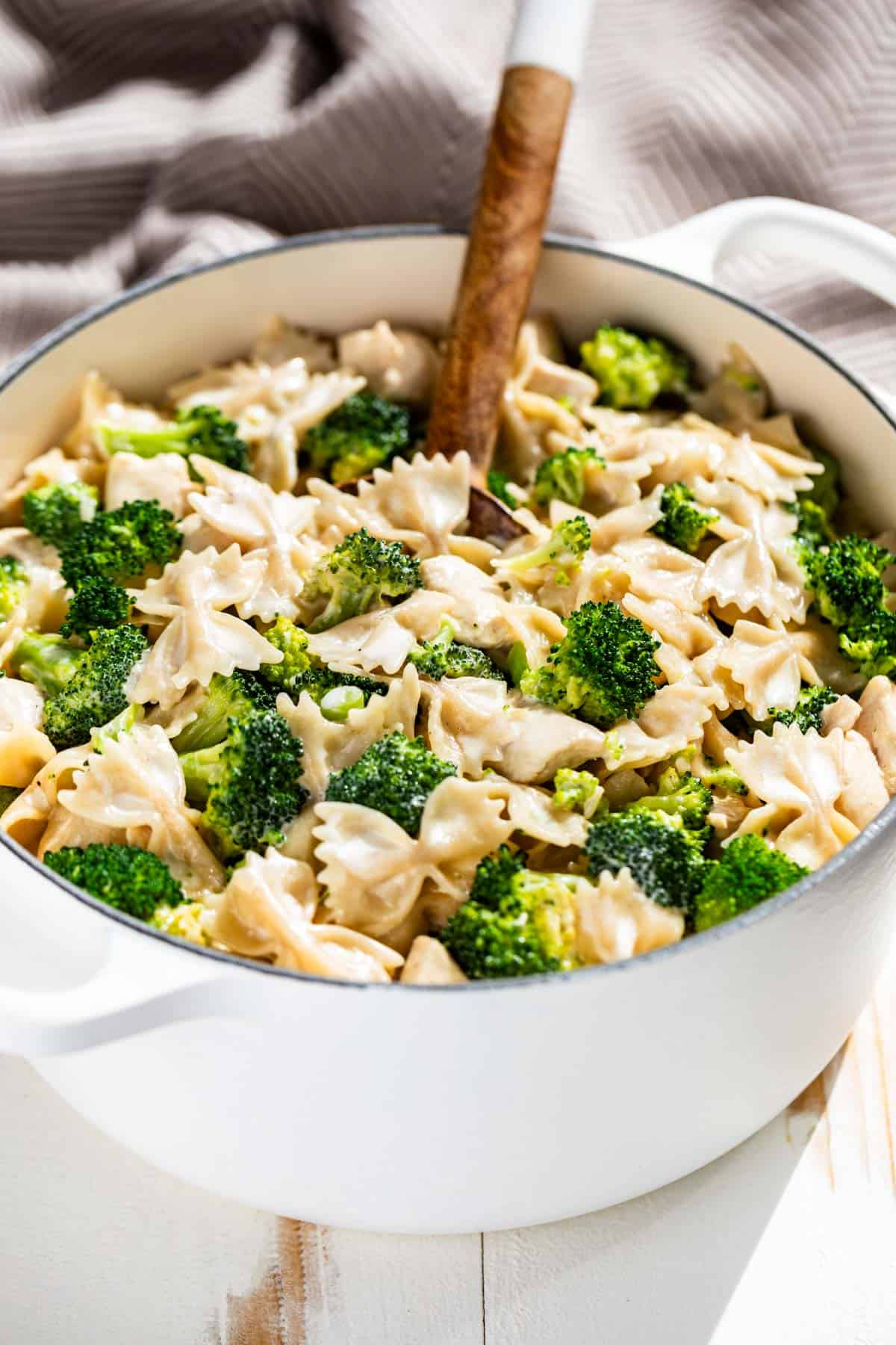 Chicken Broccoli Alfredo in a large white pot with a wood spoon in it and a brown and white striped linen in the background.