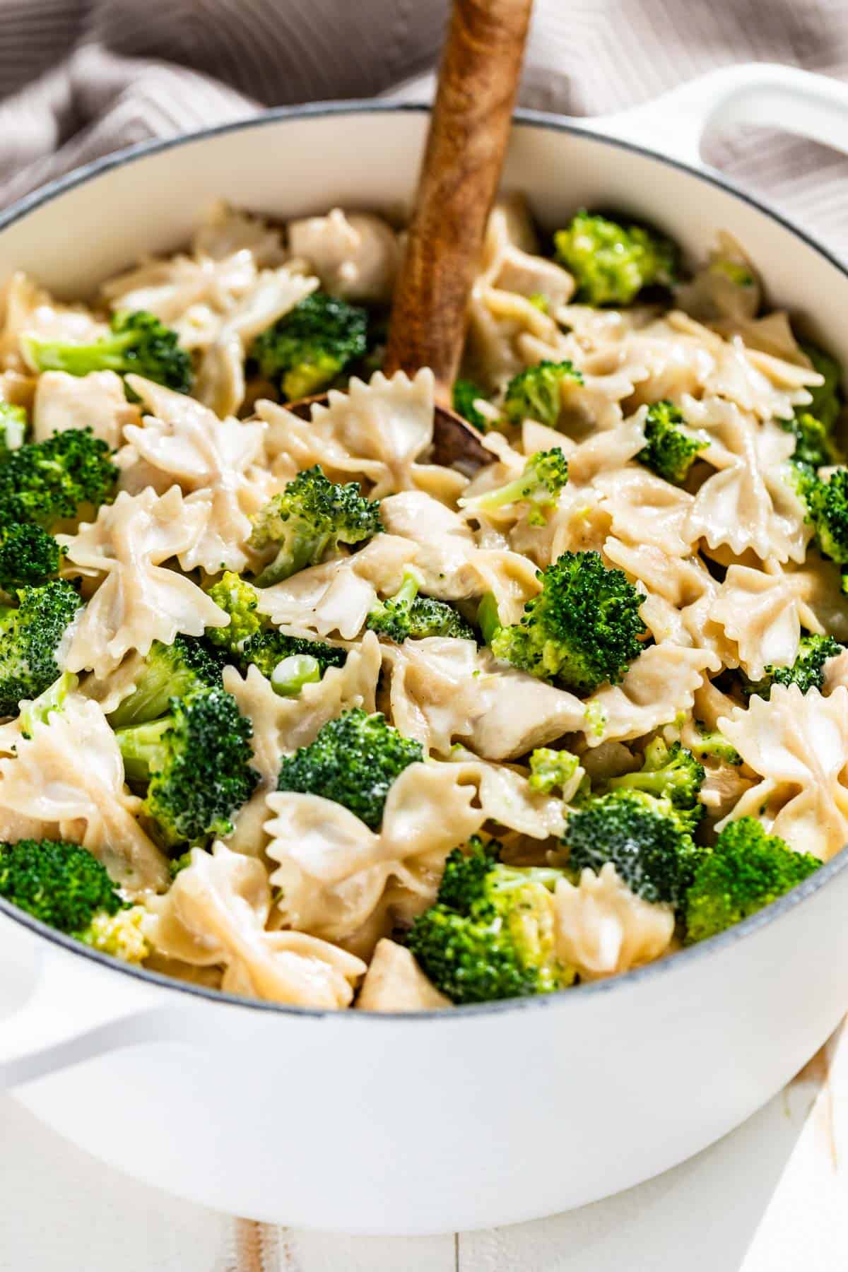 Chicken broccoli Alfredo in a large white pot with a wood spoon in it on a white background.