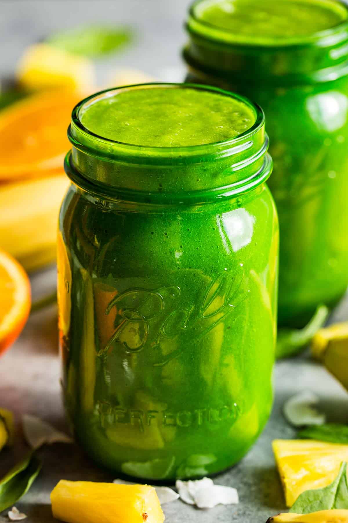 Two green jars of tropical green smoothie surrounded by pineapple pieces, flaked coconut, a banana, and sliced oranges.