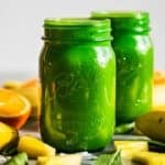 Finished Detox Island Tropical Green Smoothie in two green jars with sliced oranges, pineapple pieces, and bananas around the jars.