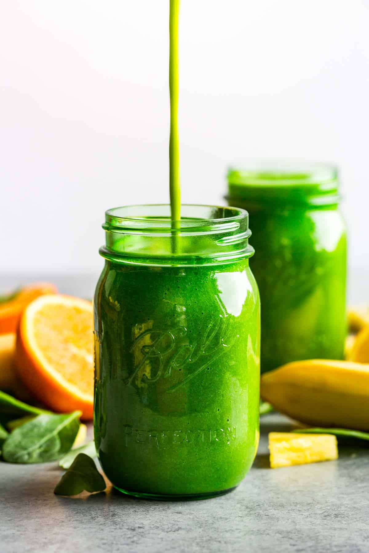 A stream of green smoothie being poured into a green jars with sliced oranges and banana around it.