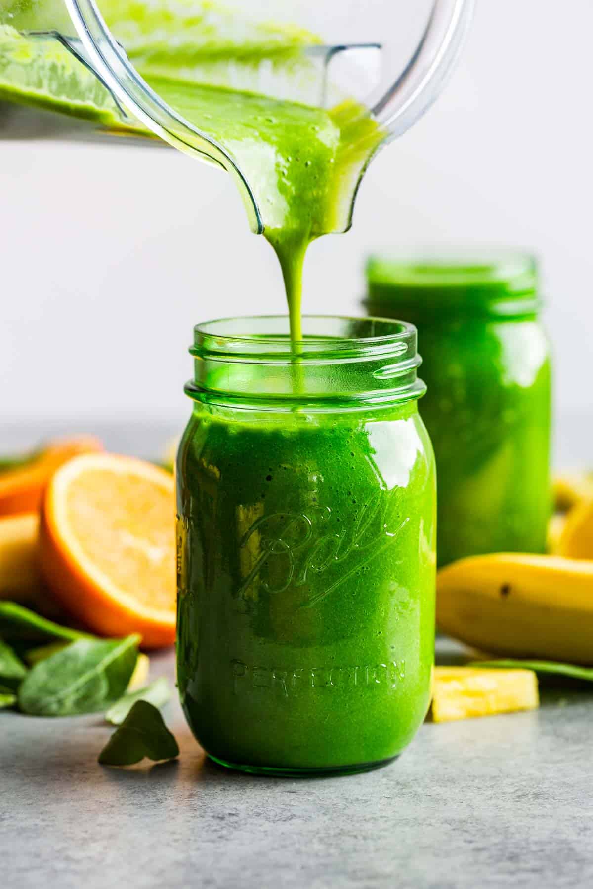A blender container pouring tropical green smoothie into two green mason jars.