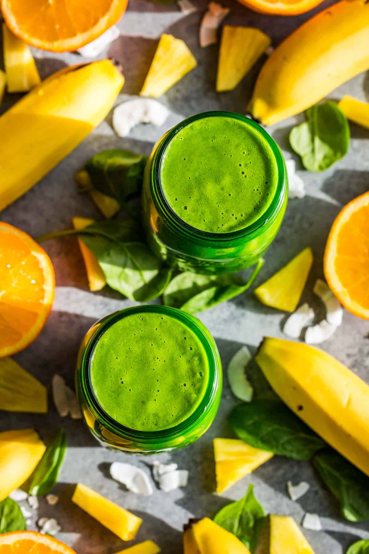 Two green jars filled with tropical green smoothie with bananas, pineapple pieces, and halved oranges around the jars.