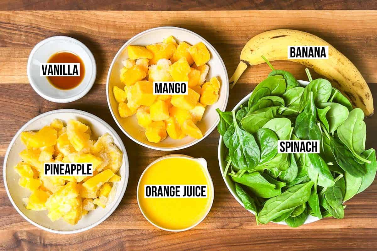 Frozen pineapple, frozen mango, orange juice, banana, vanilla, and spinach in bowls on a wood cutting board.