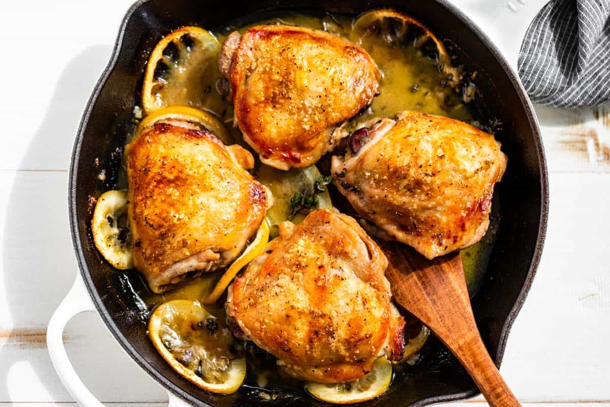 Four crispy brown chicken thighs in a large white skillet with lemon slices around them and a wood spatula scooping one chicken thigh out.