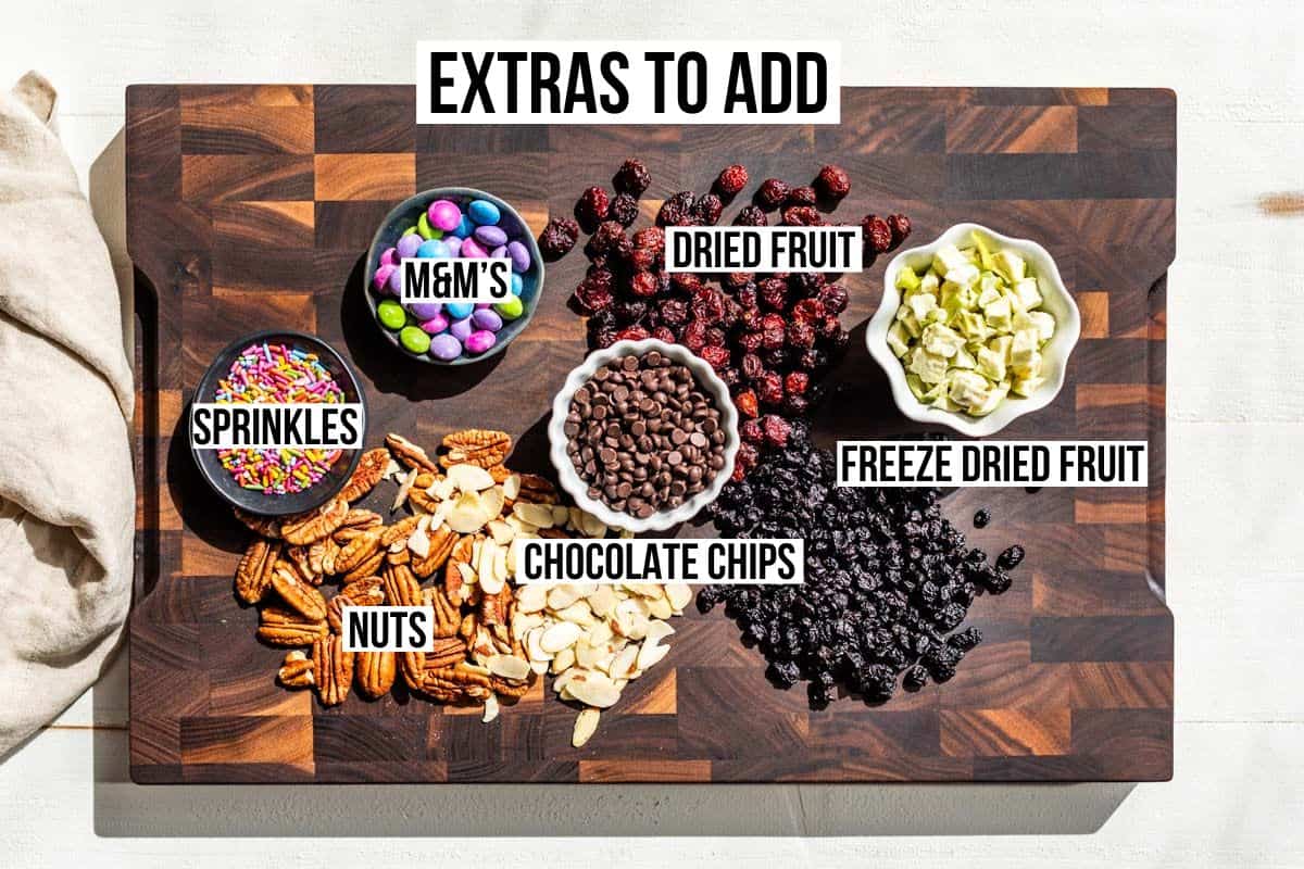 Different mix-ins for the energy bites, sprinkles, M&M's, nuts, and dried fruit on a wooden cutting board.