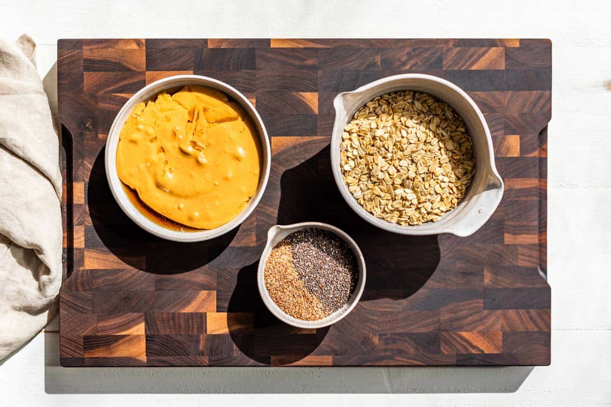 Nut butter, maple syrup, chia seeds, ground flax seeds, and oats in bowls on a wooden cutting board.