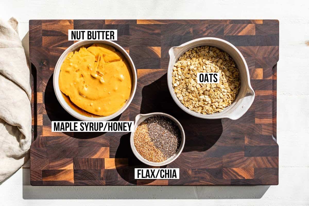 Nut butter, maple syrup, flax meal, chia seeds, and oats in pottery bowls on a wood cutting board.