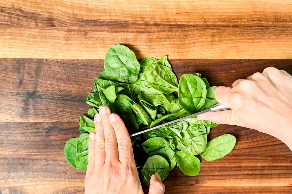 Roughly chopping the spinach on a wood cutting board with a large chef's knife.
