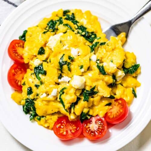 Spinach scrambled eggs with halved cherry tomatoes on the side on a white plate with a fork in the side of the eggs.
