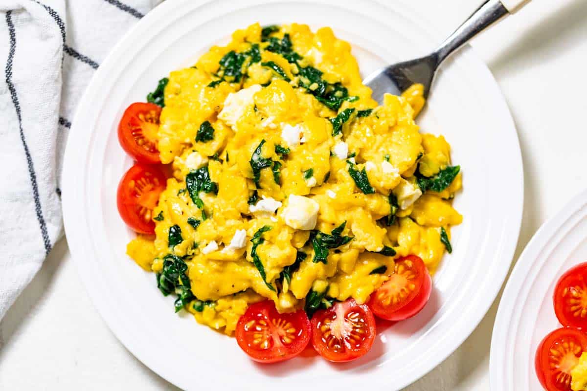 Spinach scrambled eggs with halved cherry tomatoes on the side on a white plate with a fork in the side of the eggs.