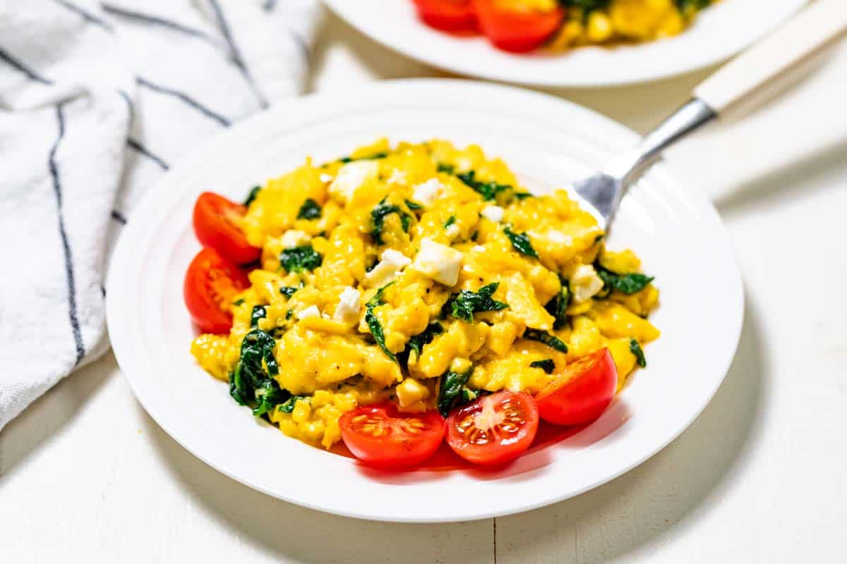 Spinach scrambled eggs with halved cherry tomatoes on the side on a white plate with a white and black striped linen on the side.