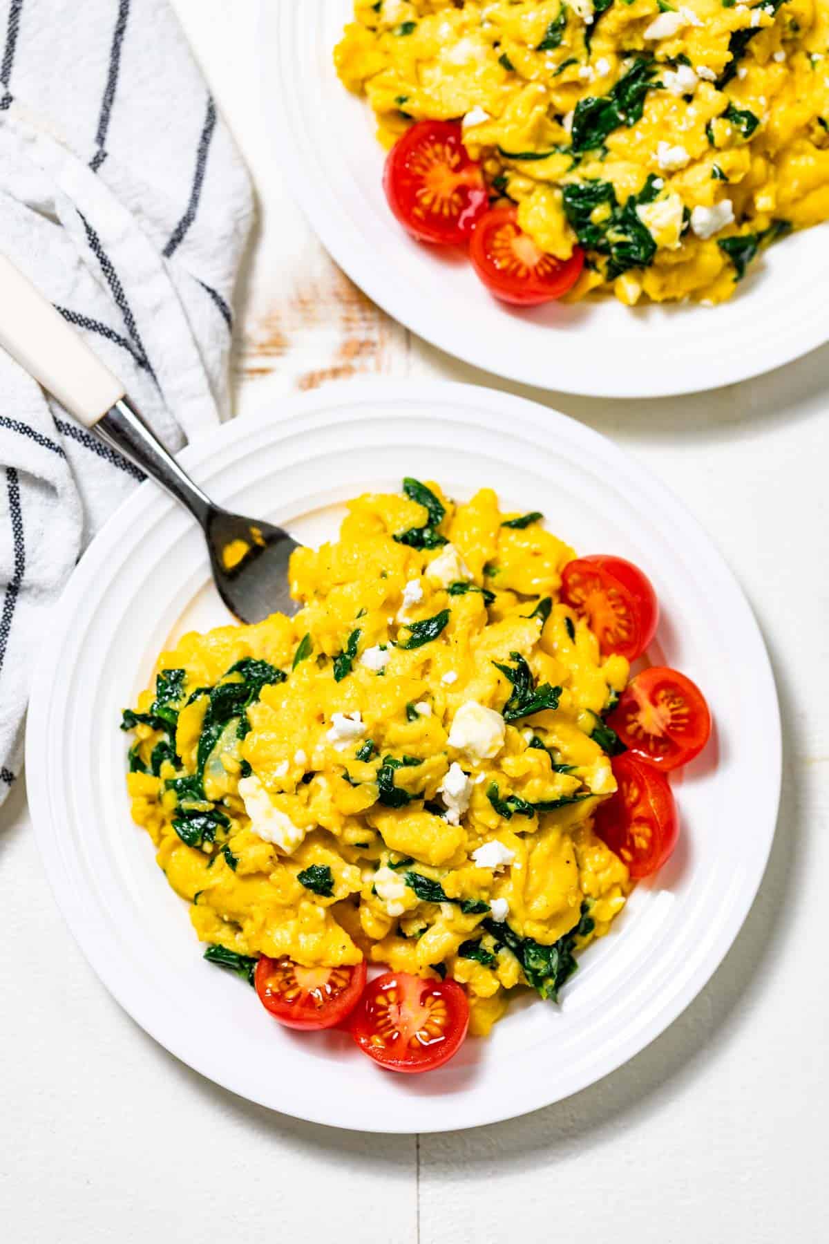 Two white plates piled up with spinach scrambled eggs with halved cherry tomatoes on the side of the plate.