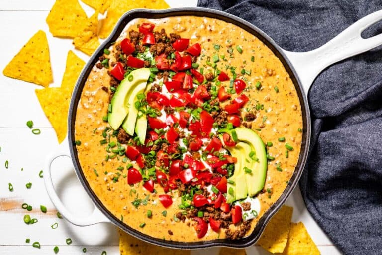 Finished Beef Queso Dip topped with diced tomatoes, green onion, cilantro, sliced avocado and sour cream surrounded by tortilla chips.