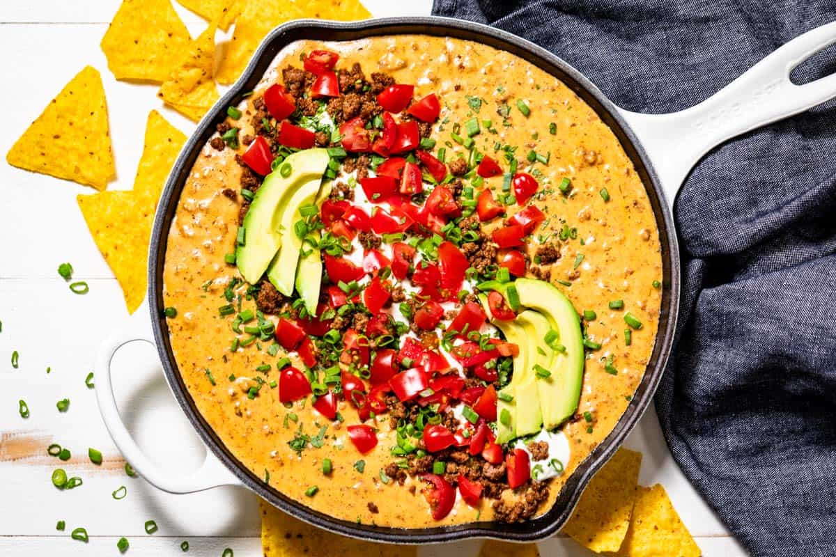 Finished Beef Queso Dip topped with diced tomatoes, green onion, cilantro, sliced avocado and sour cream surrounded by tortilla chips.