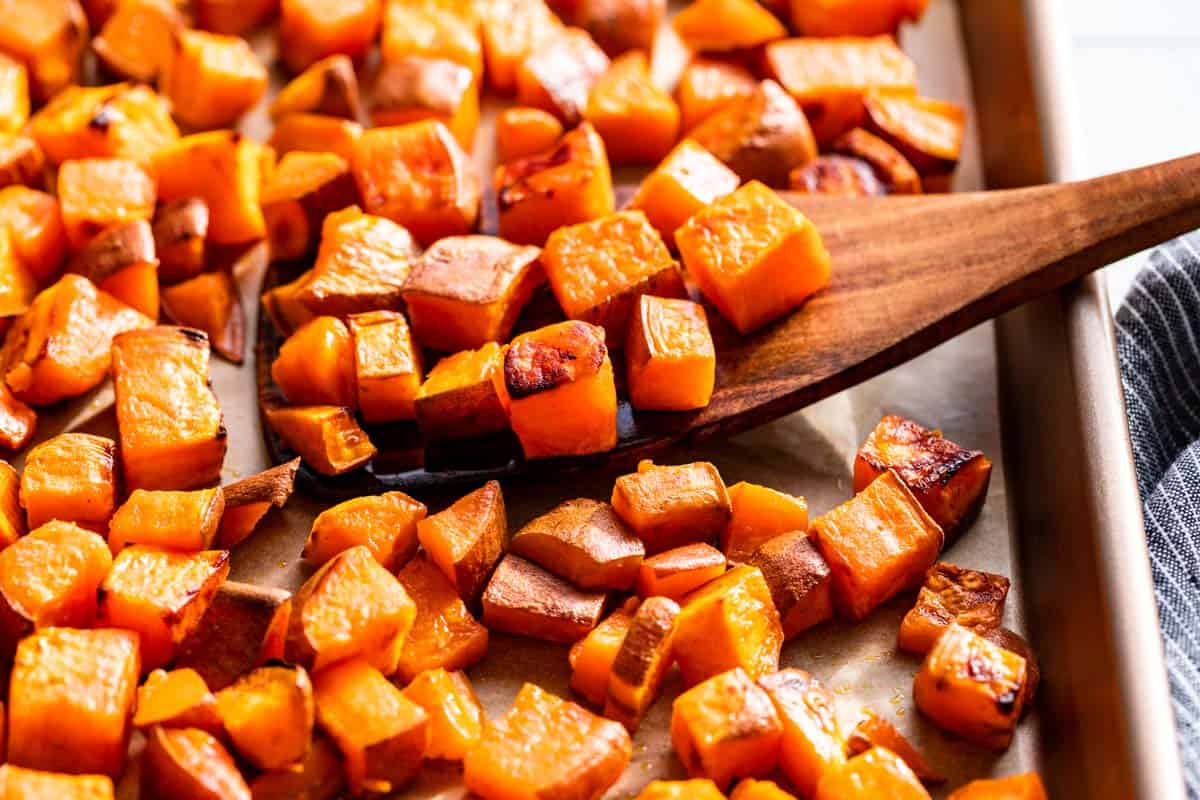 Roasted sweet potatoes on a cookie sheet with a wood spatula scooping some up.