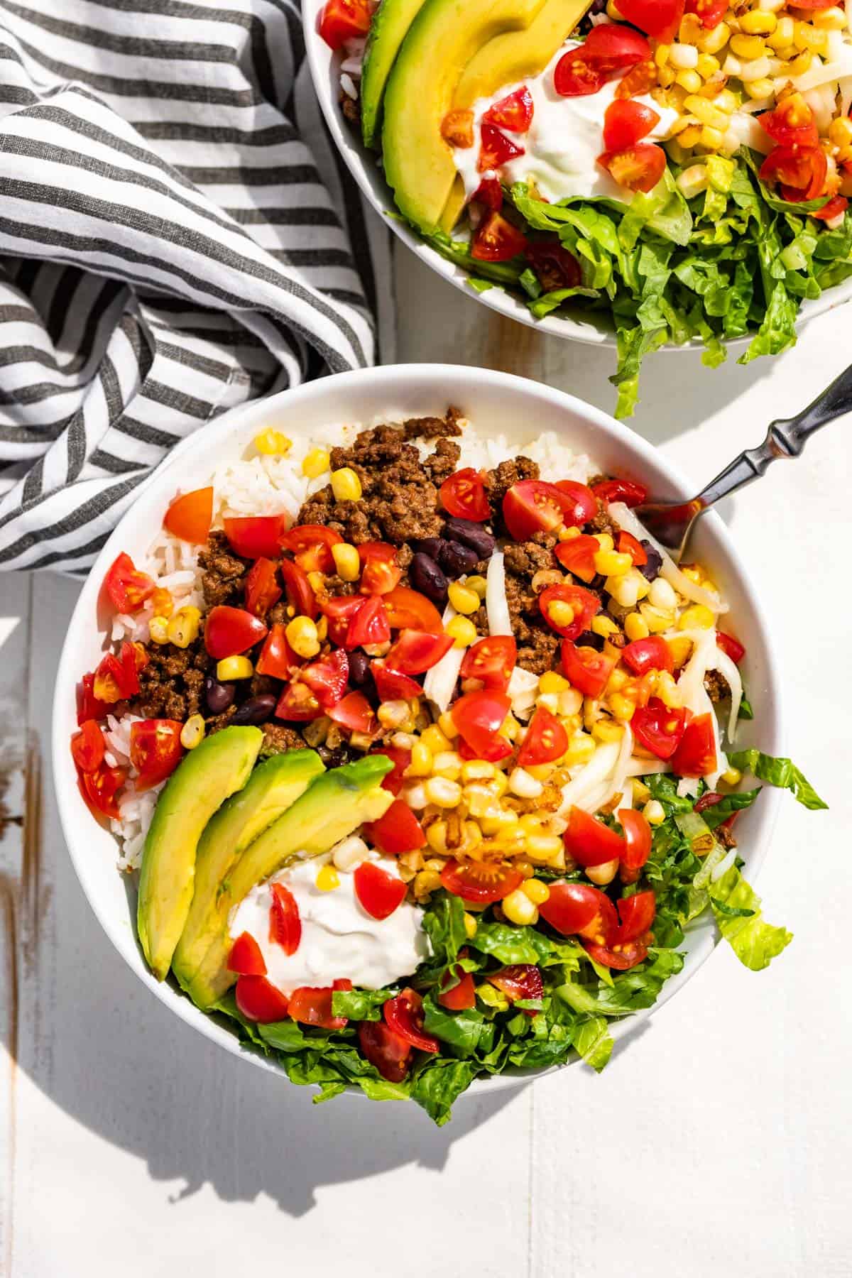 Two white bowls with rice, taco meat, chopped fresh tomatoes, sliced avocados, romaine lettuce, and corn.