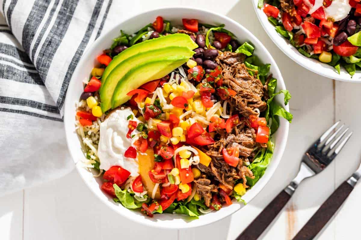Beef barbacoa in a white bowl with rice, cheese, tomatoes, avocado, corn, and sour cream.