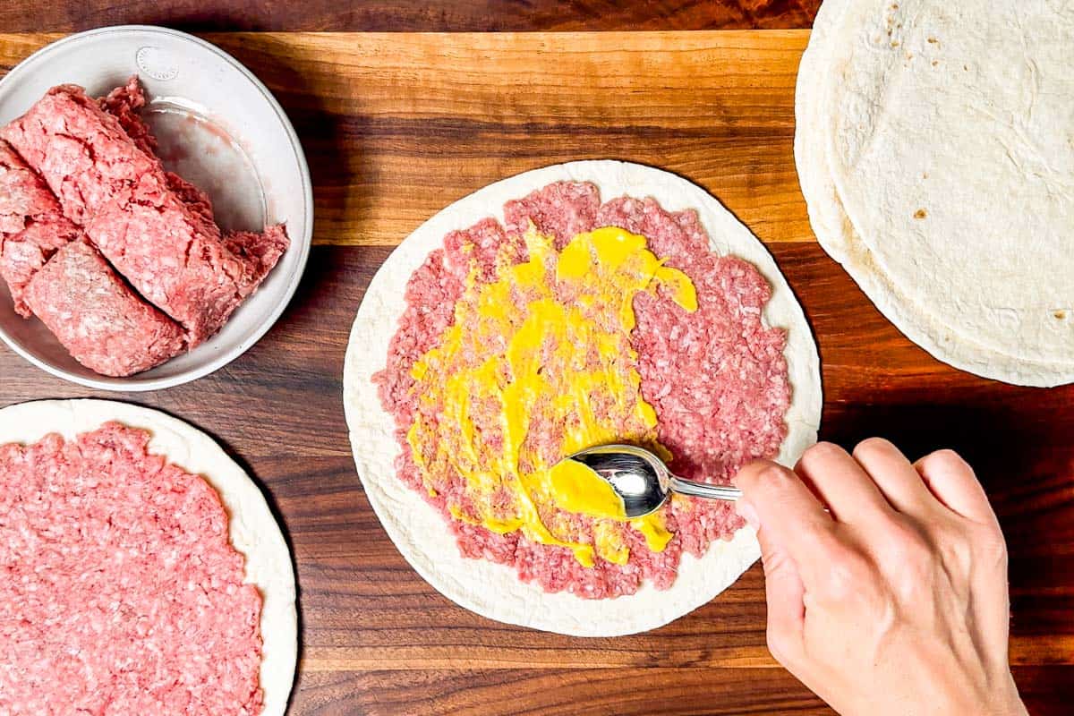 Spreading yellow mustard thinly over the ground beef smashed into the tortilla.