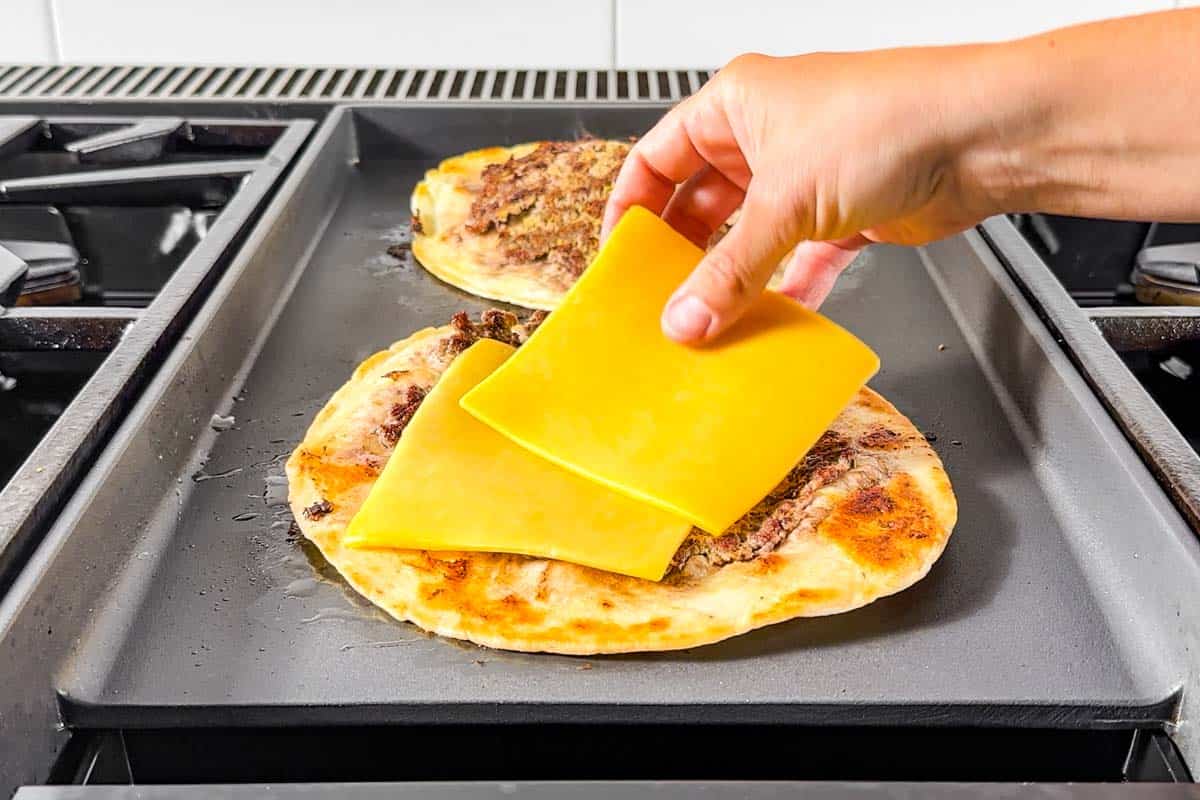 Topping the smash burger tacos with sliced cheddar cheese on a griddle.