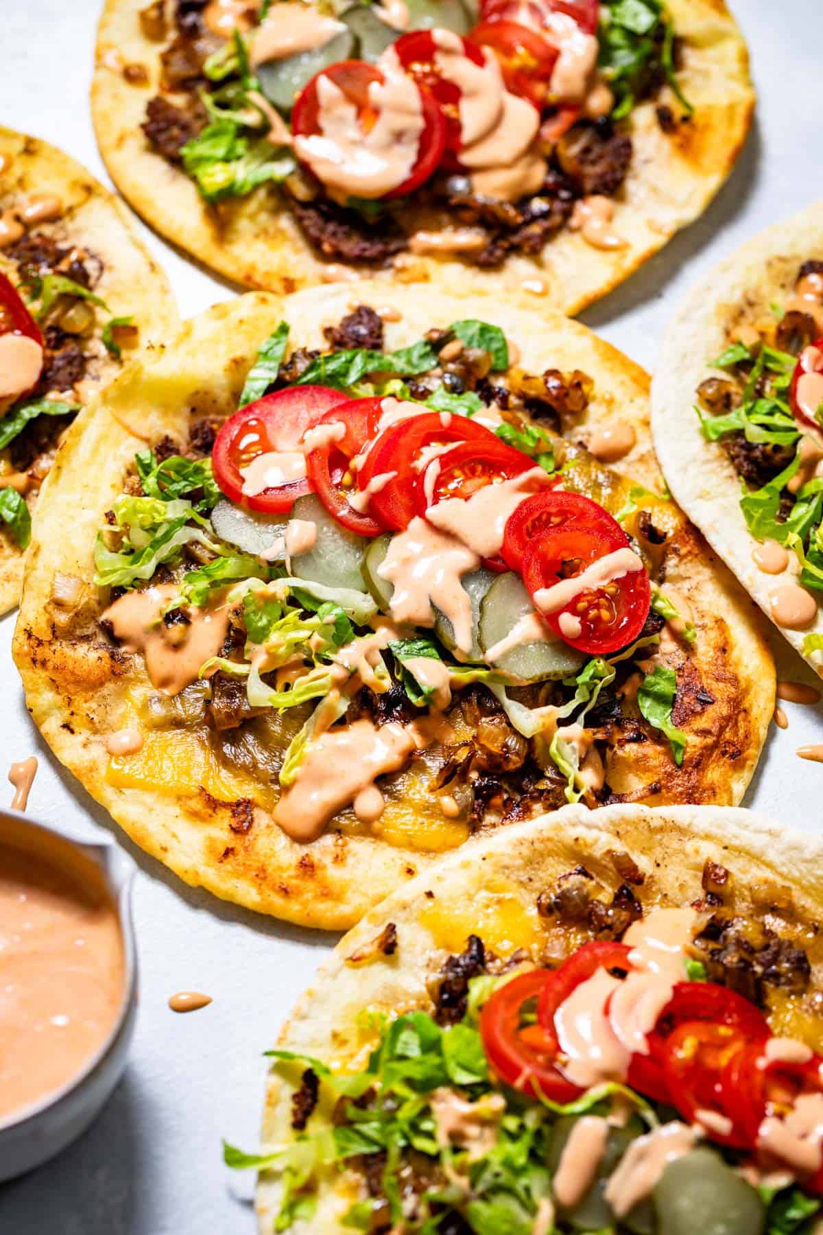 Five smash burger tacos topped with shredded lettuce, sliced tomatoes, pickles and drizzled with secret sauce with a bowl of secret sauce on the side.