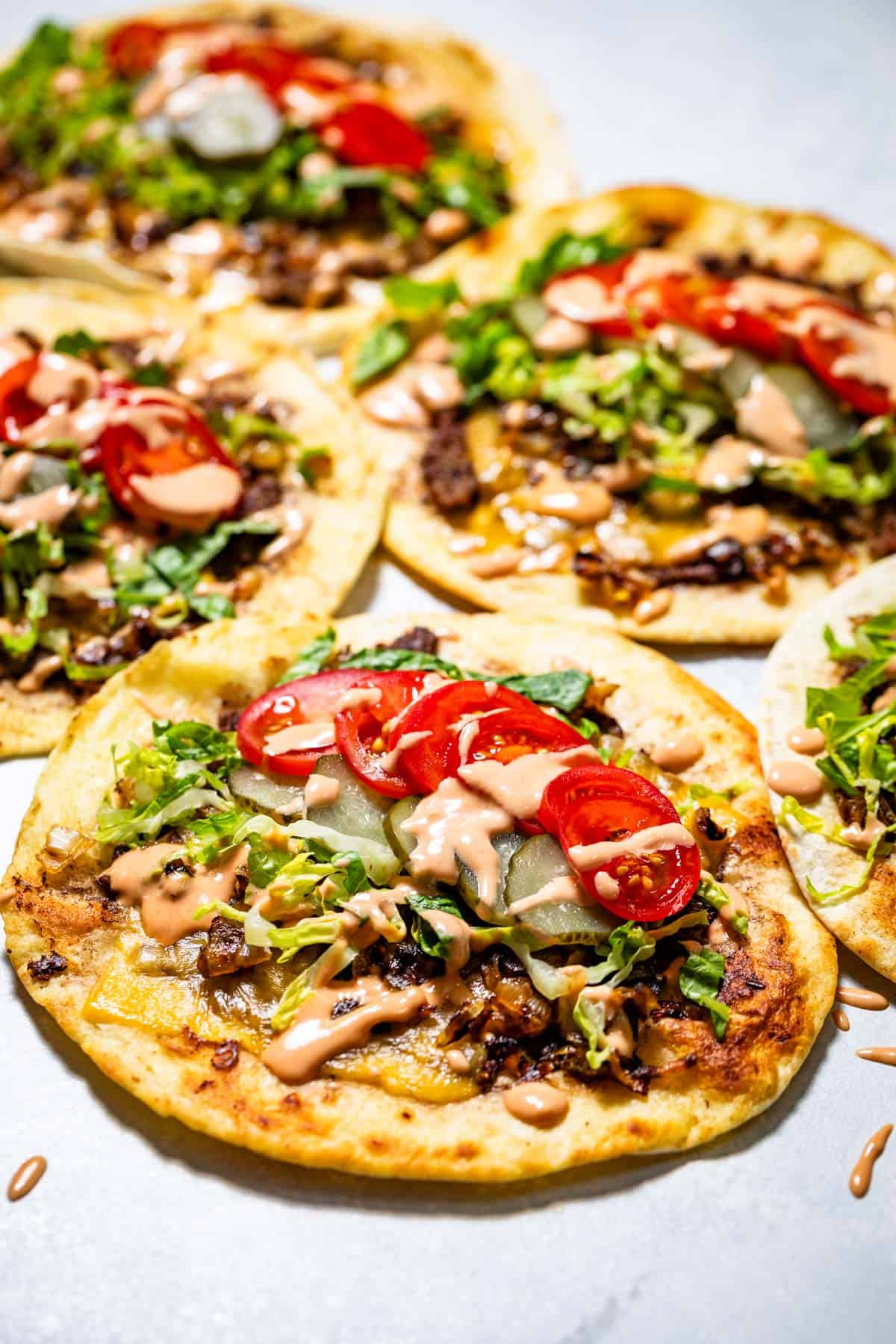 Five smash burger tacos topped with sliced tomatoes, lettuce, pickles, caramelized onions and secret sauce dressing on a blue background.