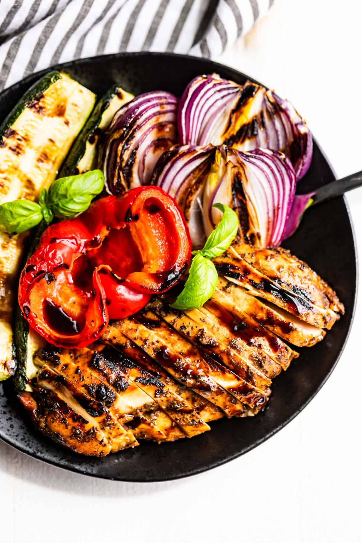 Italian balsamic grilled chicken sliced on a plate with bell pepper, red onion, and zucchini next to it.