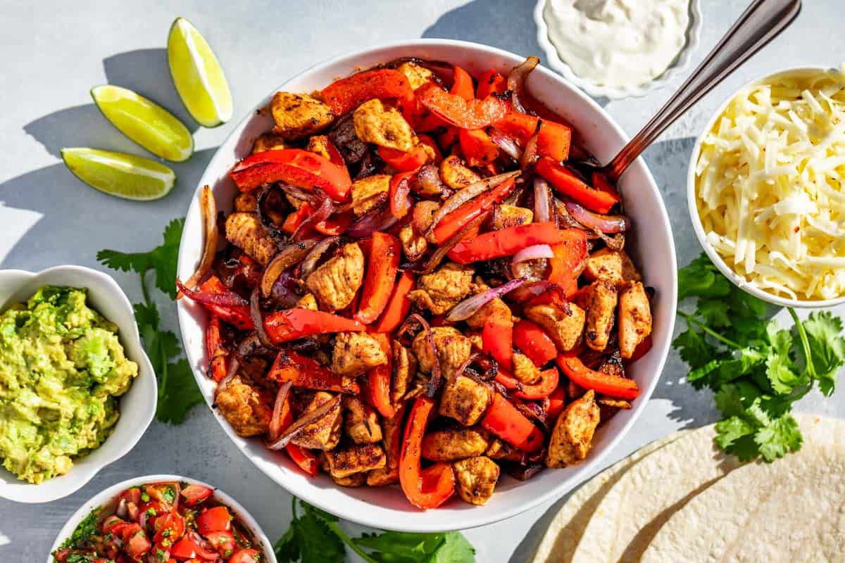 A large white bowl filled with the finished chicken fajitas surrounded by salsa, guacamole, lime wedges, cheese, cilantro, and tortillas.