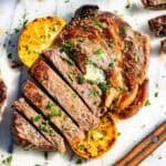 Partially sliced ribeye steak on a marble slab topped with herb butter and lemon slices with a wood knife on the side.