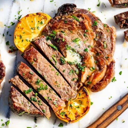 Partially sliced ribeye steak on a marble slab topped with herb butter and lemon slices with a wood knife on the side.