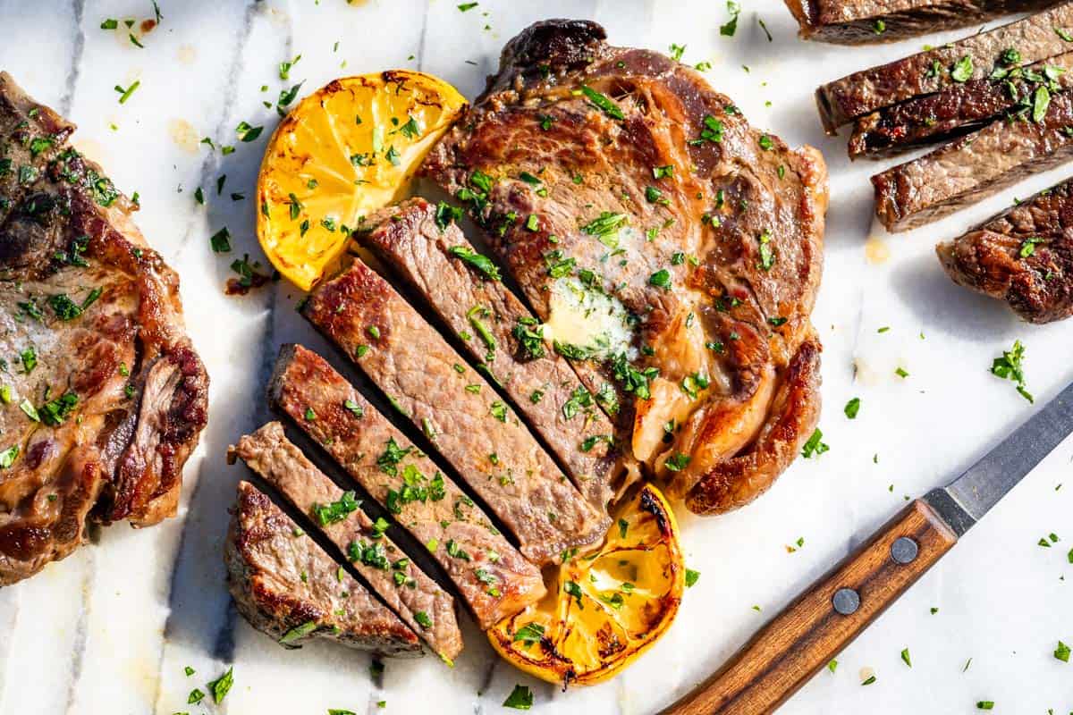 Partially sliced ribeye steak on a marble slab topped with herb butter and lemon slices with a wood knife on the side.