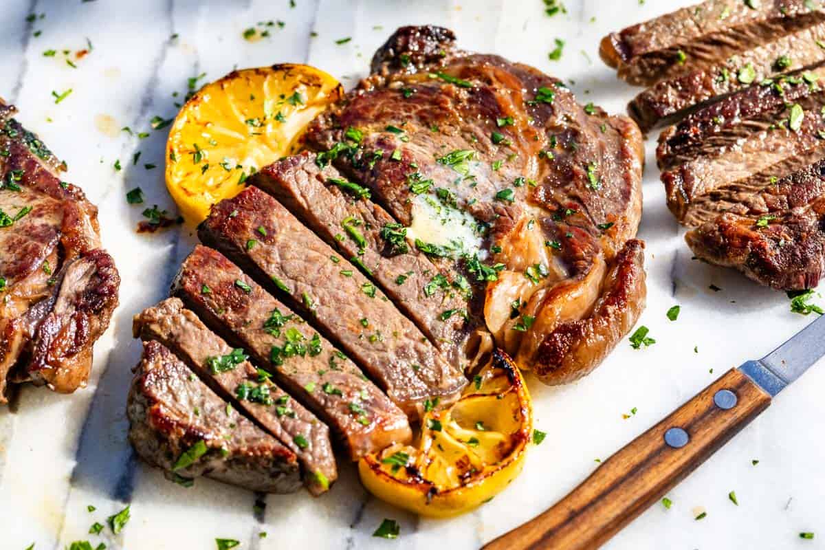 Partially sliced browned ribeye steak on a marble slab topped with herb butter and lemon slices with a wood knife on the side.