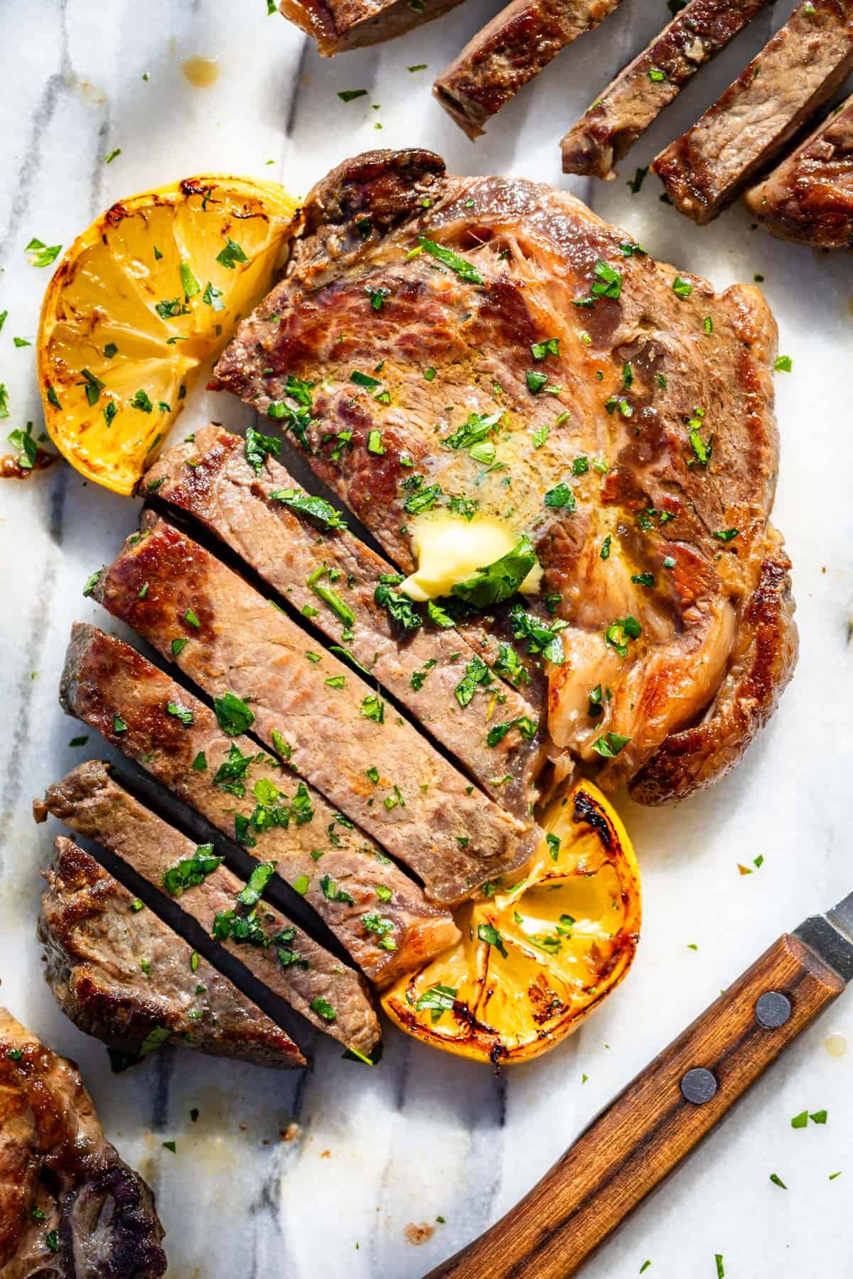 Browned sliced ribeye steak topped with herb butter and lemon slices with a wood knife on the side.