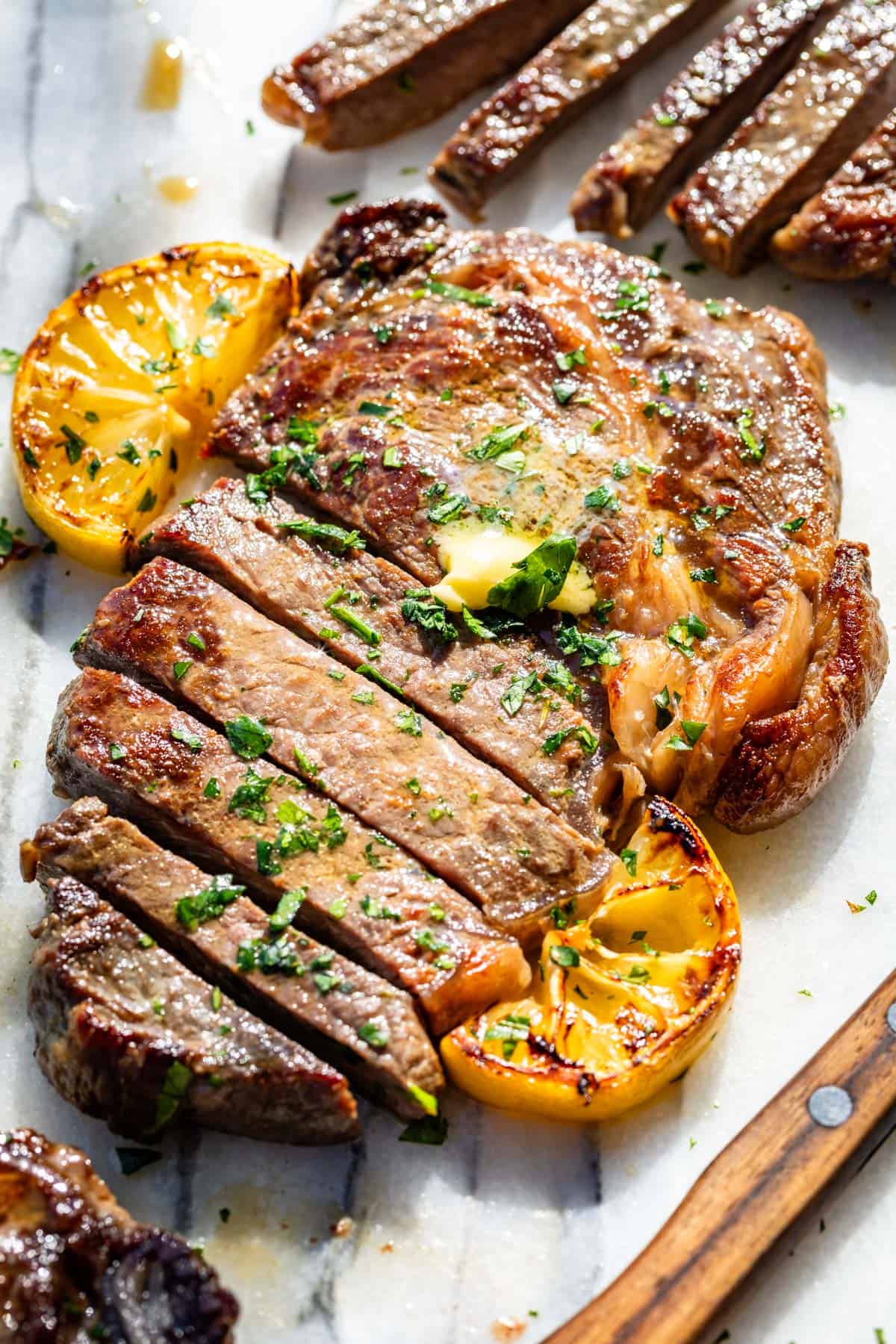Browned ribeye steak partially sliced on a marble slab topped with herb butter and lemon slices.