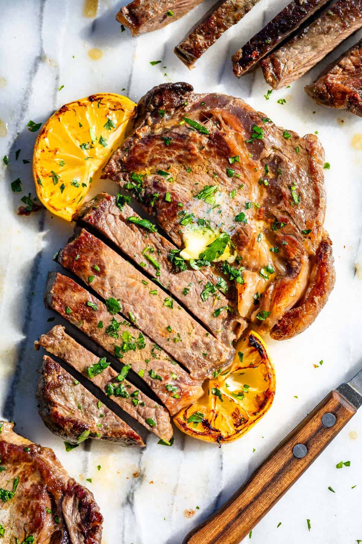 Browned ribeye steaks on marble topped with herb butter and lemon slices.