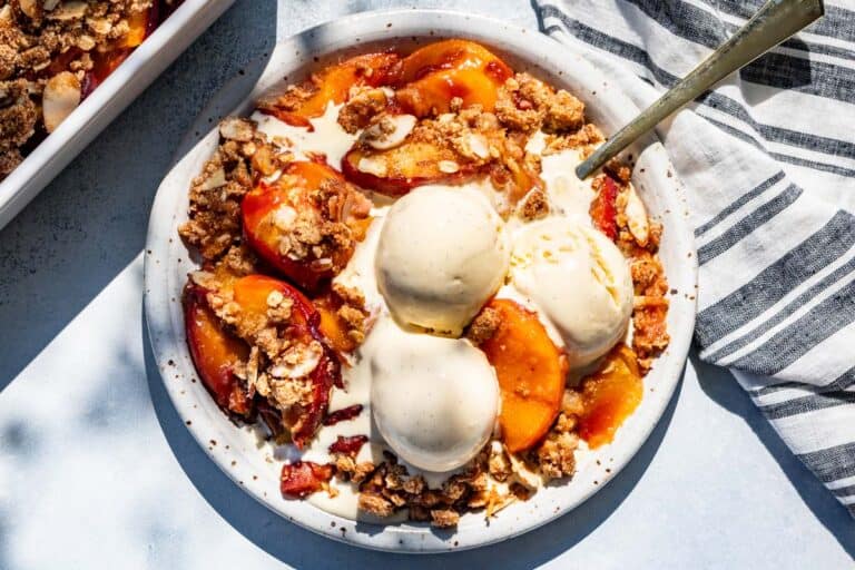 Peach crisp on a pottery plate topped with scoops of vanilla ice cream with the baking dish of peach crisp on the side.