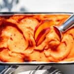 Peach sorbet in a white metal container with a black rim topped with peach slices and a ice cream scoop in the side of the sorbet.