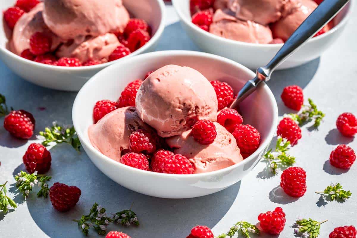 Three white bowls with scoops of raspberry ice cream topped with fresh raspberries.