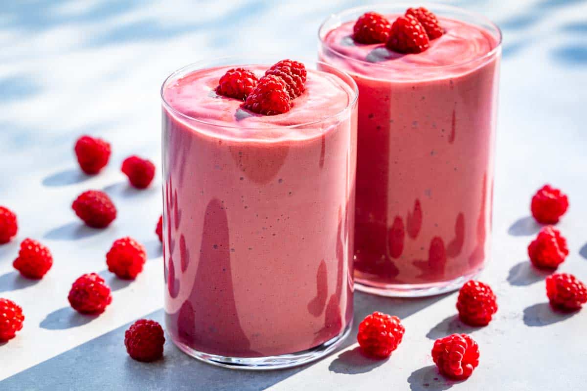 Two glasses filled with raspberry smoothie topped with raspberries and raspberries around the glasses.