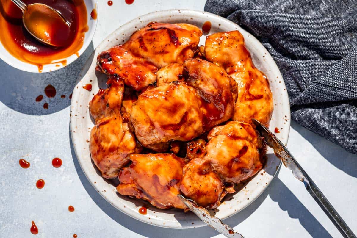 Finished smoked chicken thighs lightly glazed with BBQ sauce on a plate with metal tongs grabbing one.