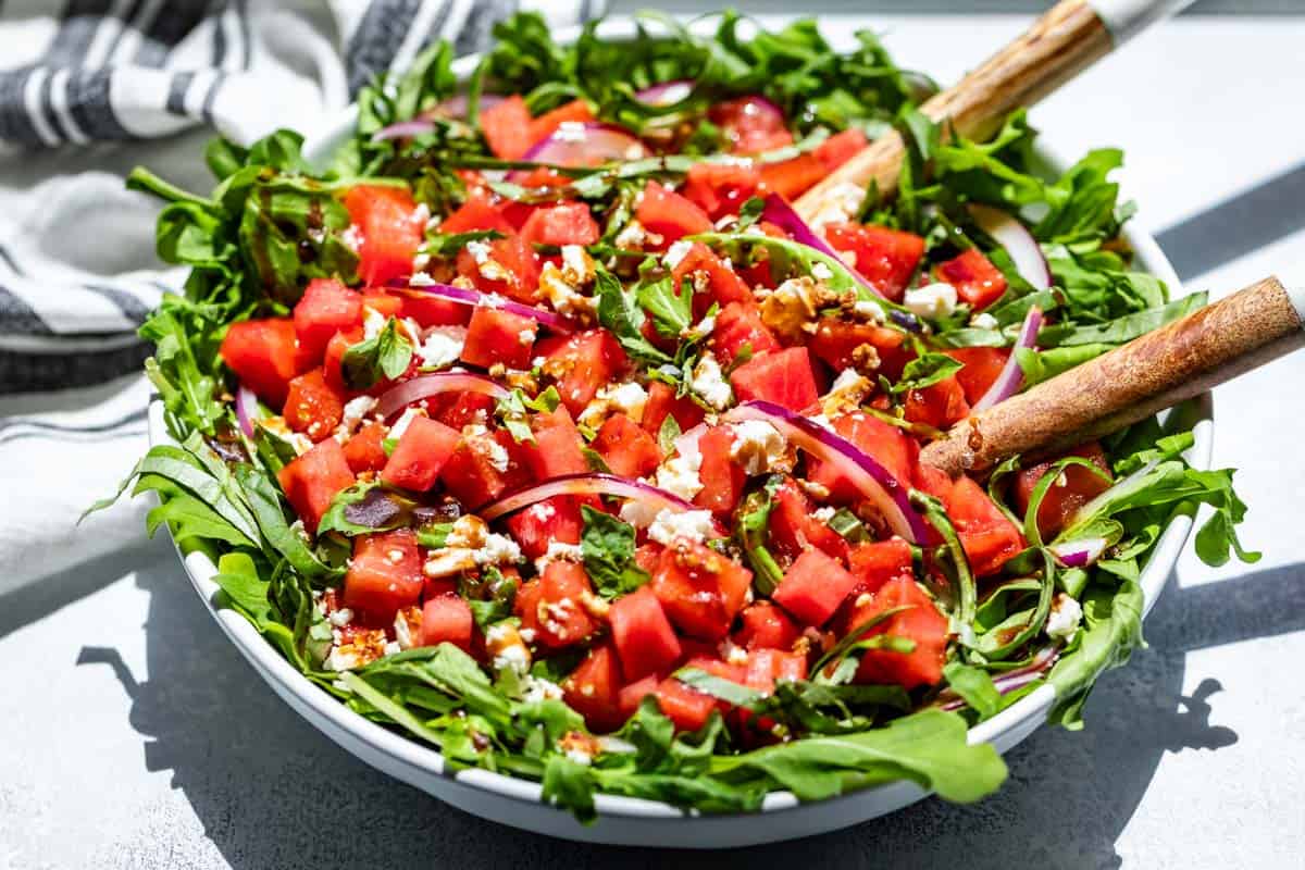 A large white bowl filled with greens topped with cubed watermelon, crumbled feta cheese, red onion and drizzled with balsamic dressing.