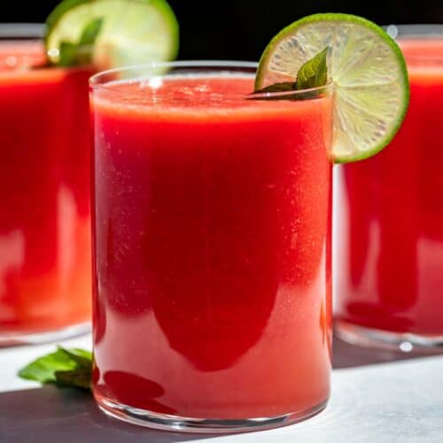 Three glasses of Watermelon Juice topped with mint sprigs and lime slices on a blue background.