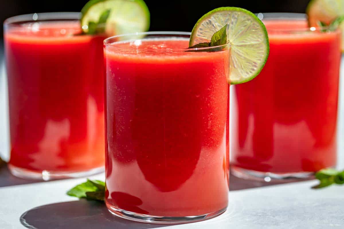 Three glasses of Watermelon Juice topped with mint sprigs and lime slices on a blue background.